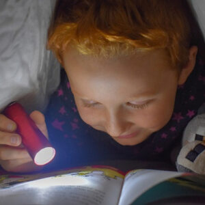 Un petit garçon tout sourire lisant en cachette sous sa couette avec une lampe torche.